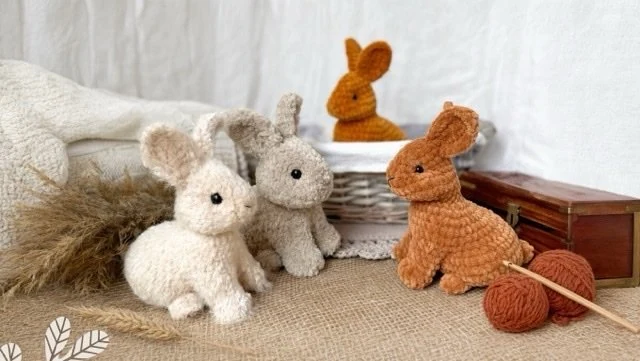 Three handmade crocheted baby rabbits displayed with yarn, a crochet hook, and a basket.