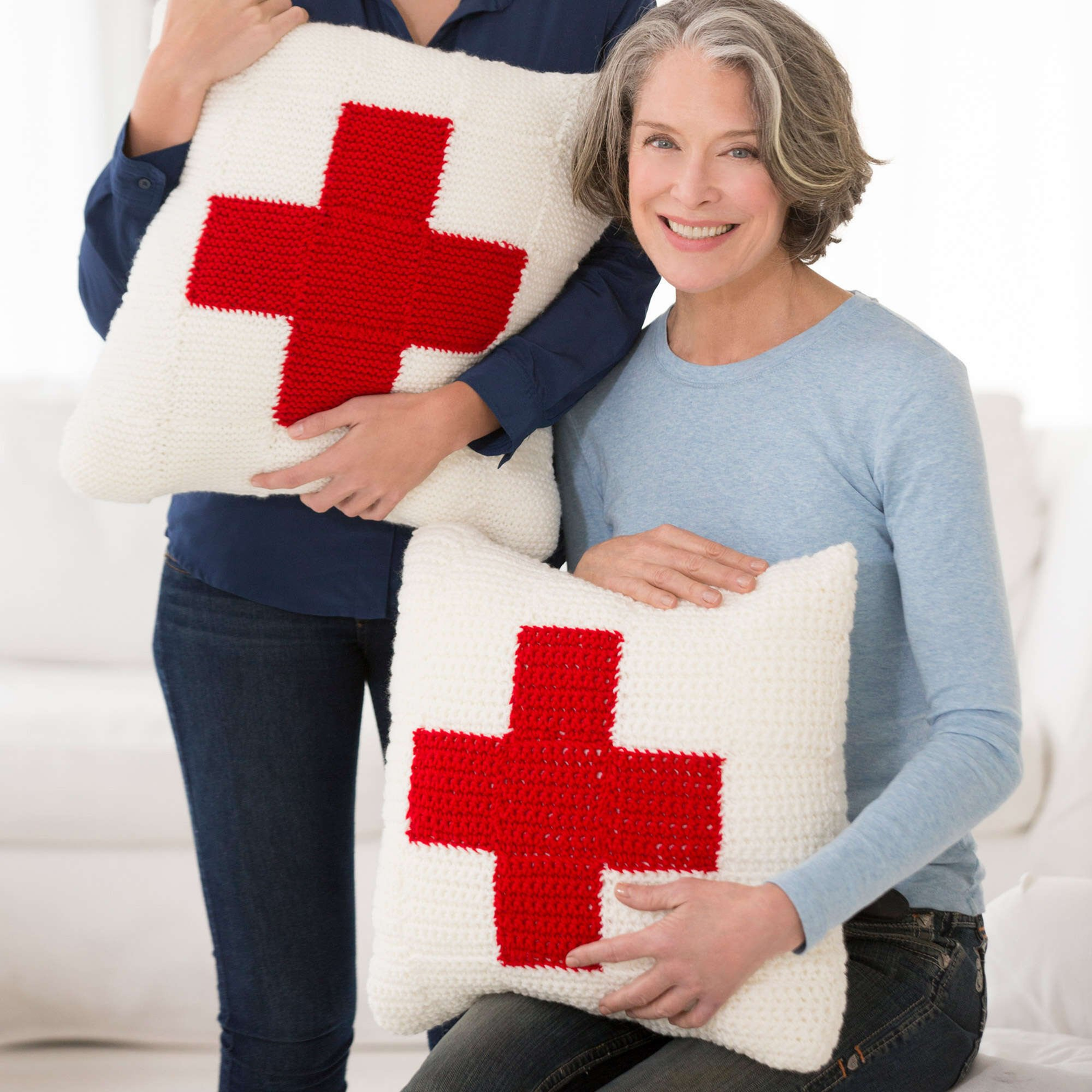 A woman holds two cream crochet pillows featuring cherry-red cross motifs.